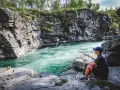 En kvinna sitter vid en forsande turkos flod omgiven av klippor. En plats för unika upplevelser i naturen.
