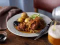 En tallrik med pannbiff, stekt lök, kokt potatis, gurka och en sked med oliver. Ett glas öl hälls upp i bakgrunden. Ett klassiskt pannbiff recept.