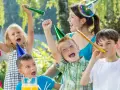 Barn firar ett 7-årskalas utomhus med ballonger, serpentiner och såpbubblor. Glada barn med partyhattar blåser i tutor.