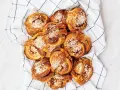 En hög med gyllene kanelbullar, toppade med pärlsocker, ligger på en vit handduk med blå rutor. Camilla Hamid skulle vara stolt över dessa kanelbullar.