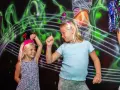 Två glada barn med diadem har roliga lekar inomhus. De dansar framför en neonfärgad bakgrund.