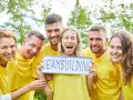 En grupp glada människor i gula tröjor håller upp en skylt med texten "TEAMBUILDING". De ser ut att ha roliga uppdrag i grupp.