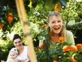 Två glada kvinnor och en bebis njuter av en solig dag utomhus, kanske under en tipspromenad med utmaningar för vuxna bland blommor och grönska.