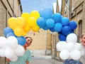 Två glada personer gömmer sig bakom en båge av gula, blå och vita ballonger. Perfekt för studentmottagning lekar!