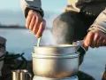En person lagar mat utomhus, ångande kastruller och en mugg i metall antyder en lyxig matupplevelse i naturen.