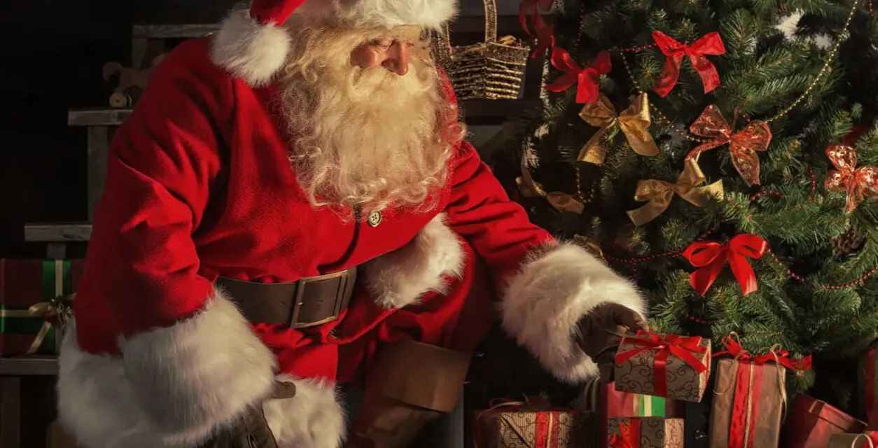 Jultomten, med sitt vita skägg och röda dräkt, sorterar julklappar vid ett dekorerat träd. Här är fakta om jultomten.