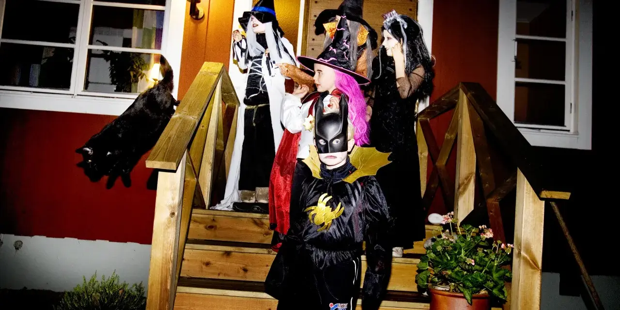 Barn utklädda till häxor och Batman förbereder sig för halloween helg. En svart katt smyger på trappan.