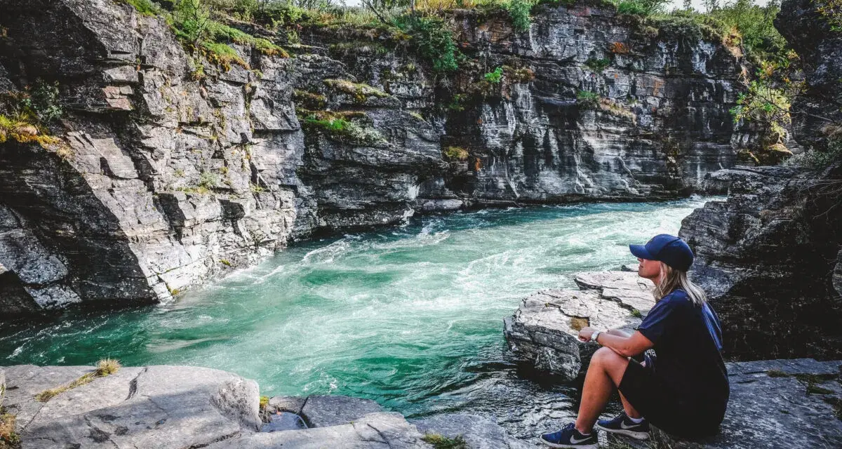 En kvinna sitter vid en forsande turkos flod omgiven av klippor. En plats för unika upplevelser i naturen.