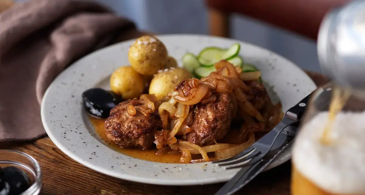 En tallrik med pannbiff, stekt lök, kokt potatis, gurka och en sked med oliver. Ett glas öl hälls upp i bakgrunden. Ett klassiskt pannbiff recept.