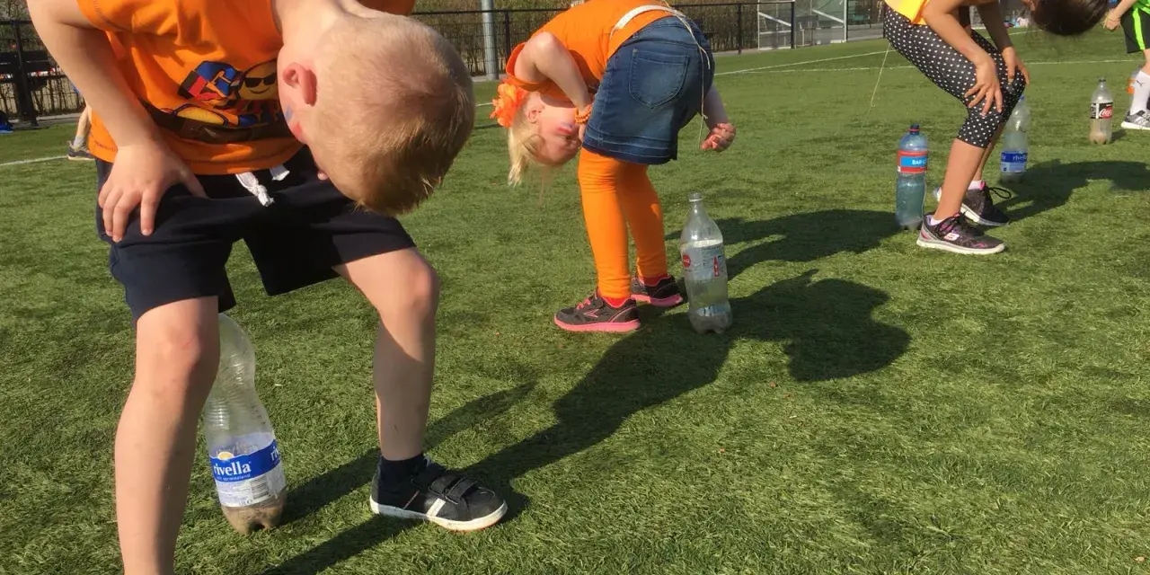 Barn i orange kläder böjer sig framåt för att försöka fånga en flaska med munnen. En del av kalas lekarna.