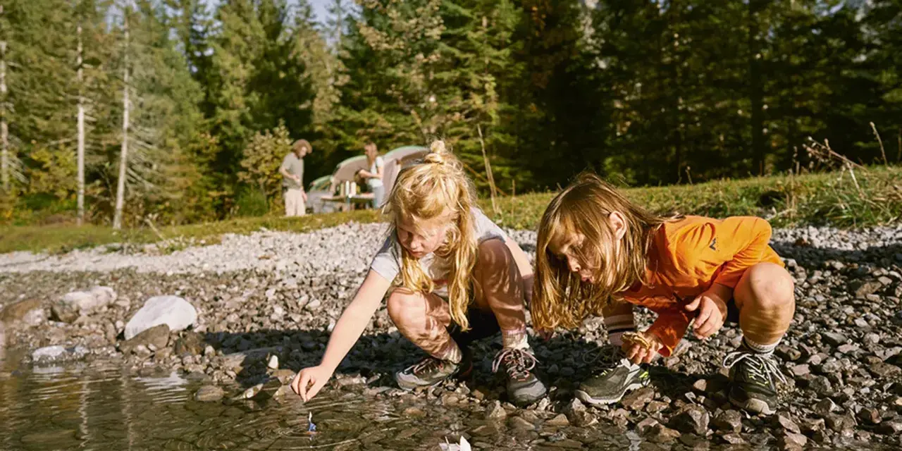 Två barn leker med pappersbåtar vid en sjö, kanske en del av roliga femkampsgrenar. I bakgrunden syns ett tält och träd.