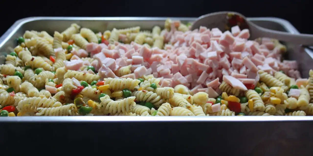 Stor ugnsform fylld med pastasallad med kassler, grönsaker och en sked.