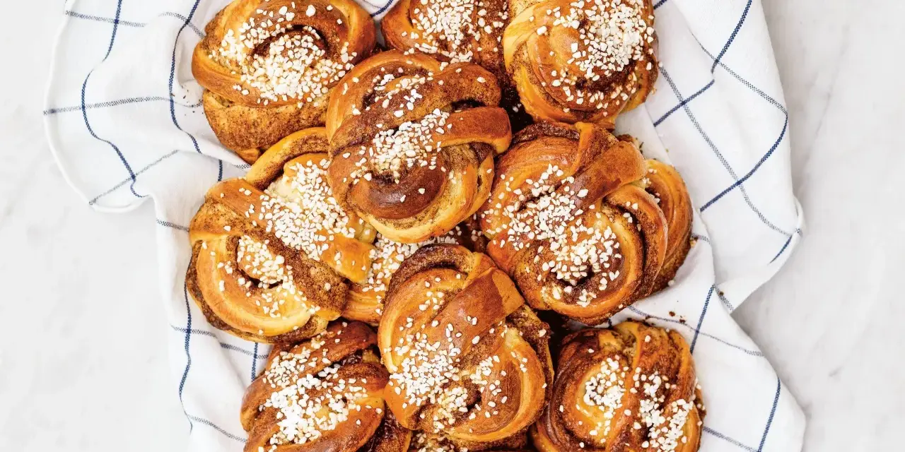 En hög med gyllene kanelbullar, toppade med pärlsocker, ligger på en vit handduk med blå rutor. Camilla Hamid skulle vara stolt över dessa kanelbullar.