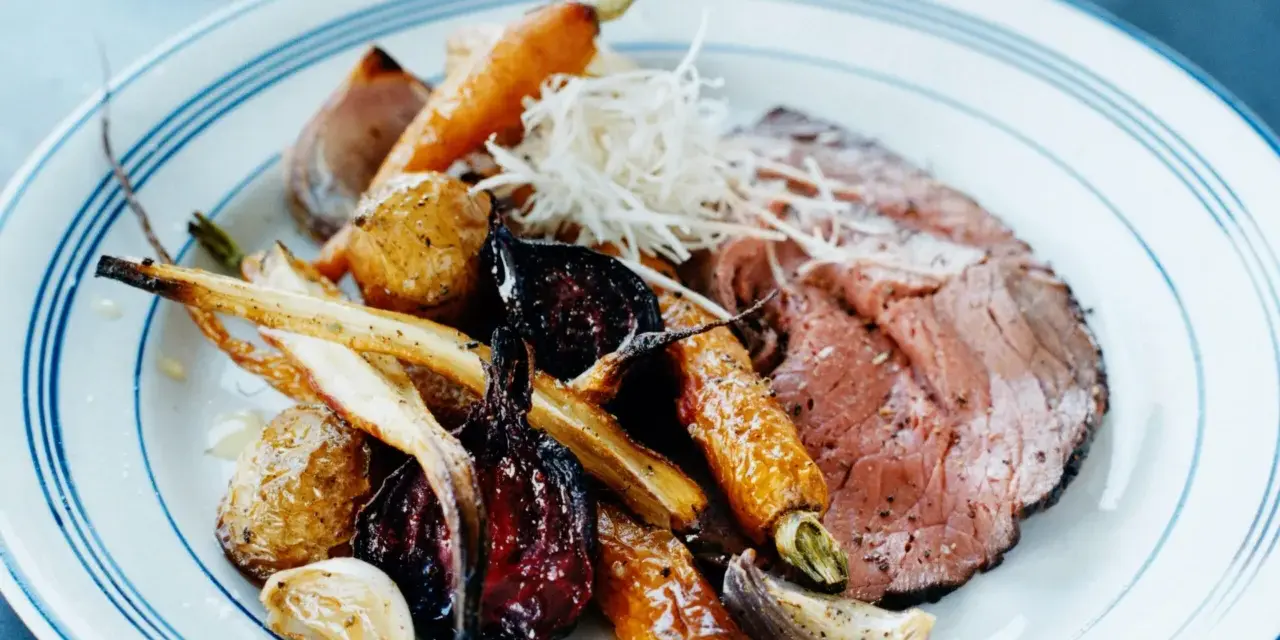 En tallrik med skivad rostbiff, rostade rotfrukter som morötter och palsternacka, samt vitlök. Ett perfekt rostbiff recept för en festmåltid.