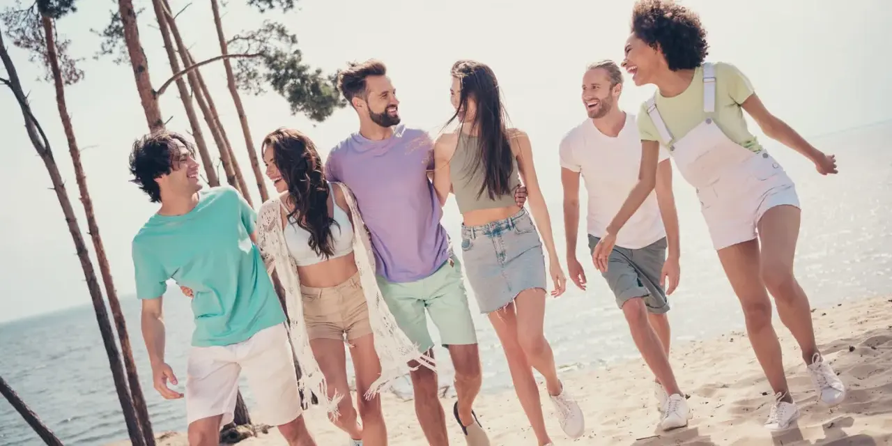 En grupp glada vänner på stranden, en riktig tipspromenad med twist. De skrattar och går längs vattnet bland tallar.