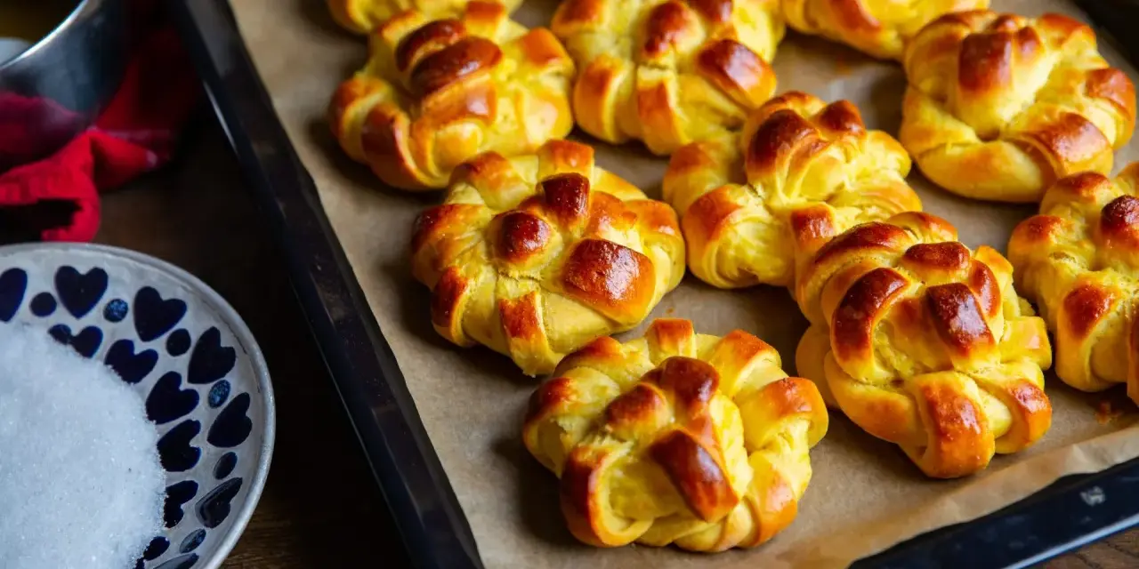 Nybakade lussebullar med kesella, gyllene och redo att avnjutas. En skål med socker och en röd duk anas i bakgrunden.