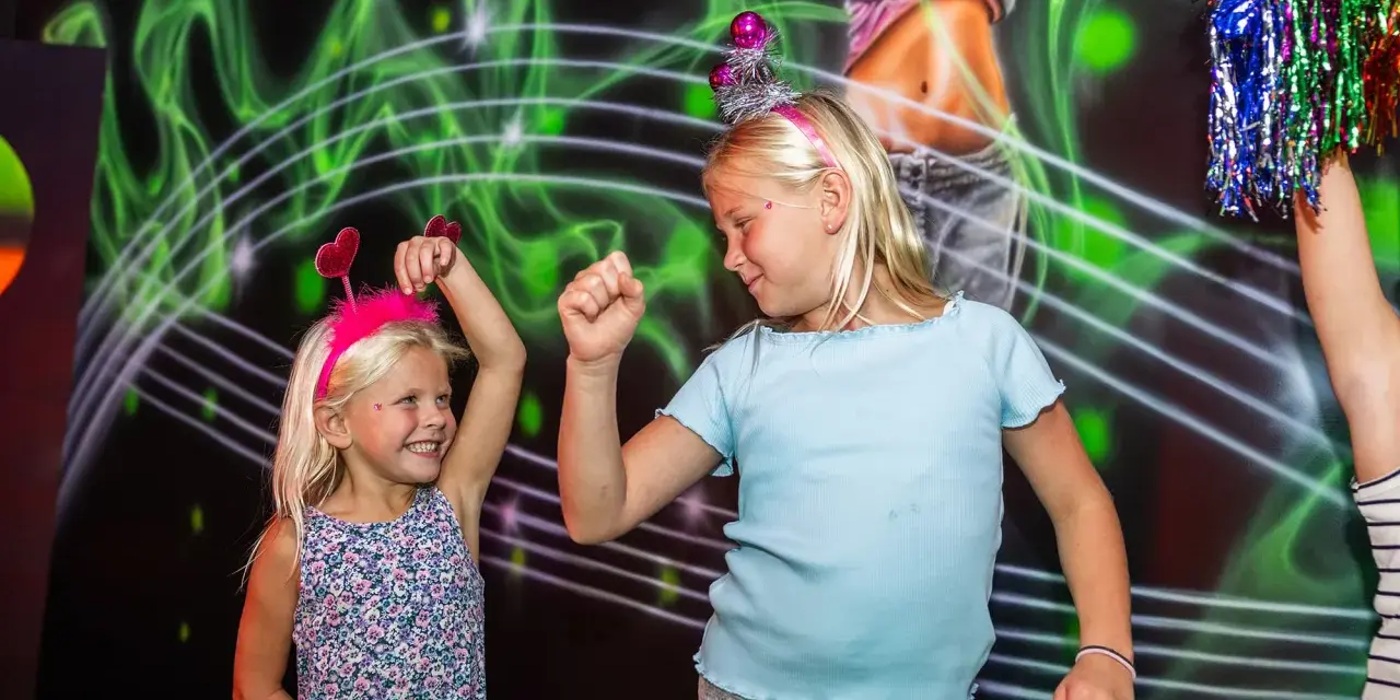 Två glada barn med diadem har roliga lekar inomhus. De dansar framför en neonfärgad bakgrund.