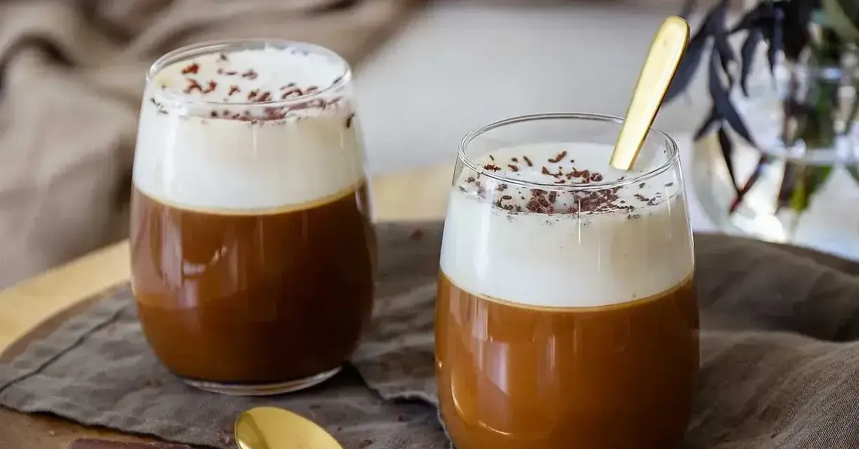 Två glas med krämig kaffe Karlsson, toppad med vispad grädde och chokladströssel. En guldig sked ligger i det ena glaset.
