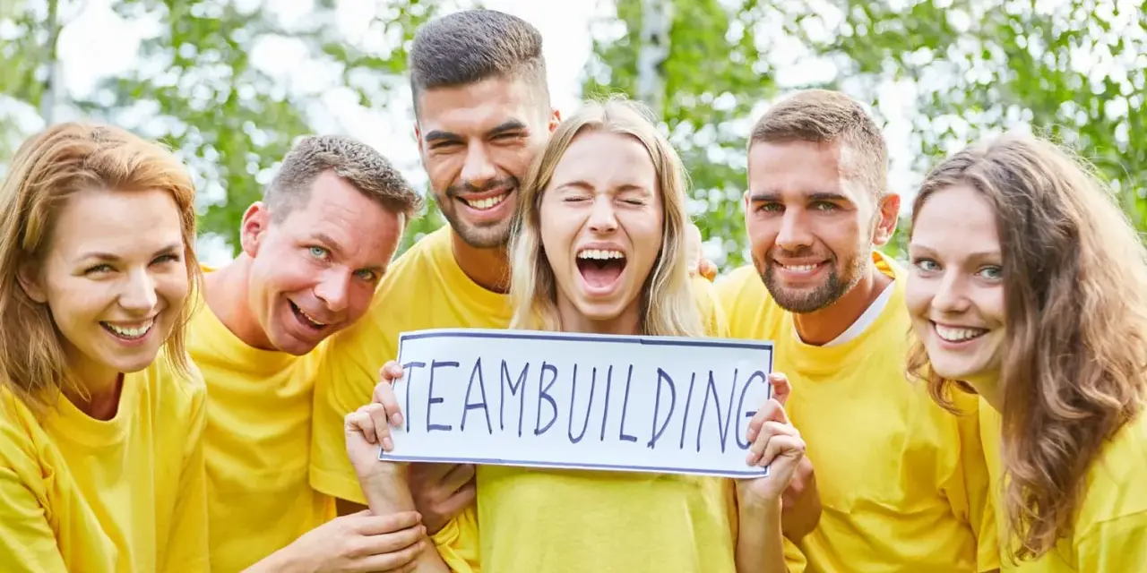 En grupp glada människor i gula tröjor håller upp en skylt med texten "TEAMBUILDING". De ser ut att ha roliga uppdrag i grupp.