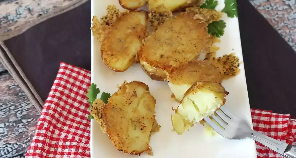 Krispiga, gyllene smashed potatis på en vit tallrik, garnerade med persilja. En gaffel visar en mjuk insida.