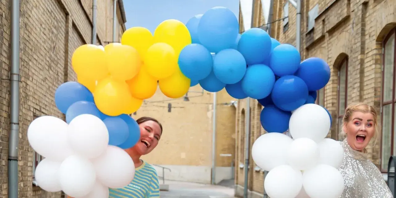 Två glada personer gömmer sig bakom en båge av gula, blå och vita ballonger. Perfekt för studentmottagning lekar!