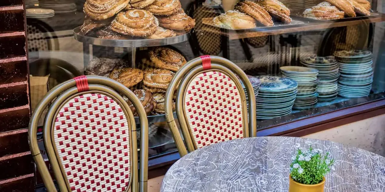 Två stolar med flätad sits och ryggstöd framför ett skyltfönster med kanelbullar och andra bakverk. Ett annorlunda café med charmig inredning.