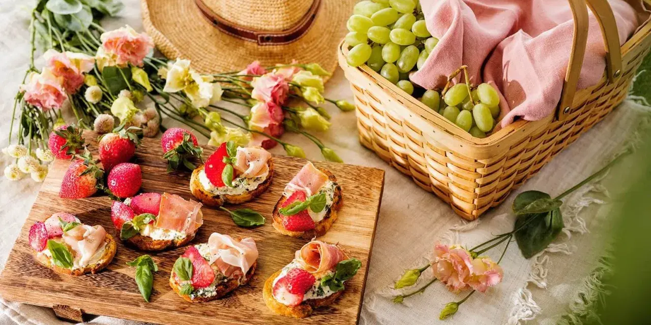 En härlig picknick med bruschetta toppad med jordgubbar och parmaskinka, färska jordgubbar, vindruvor i korg och en stråhatt.