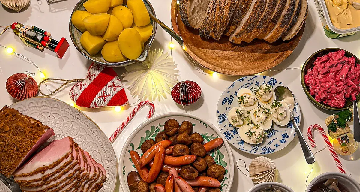 Julbord med jullekar vid bordet: skinka, köttbullar, prinskorv, sill, ägg och bröd. Julpynt och ljusslingor skapar stämning.