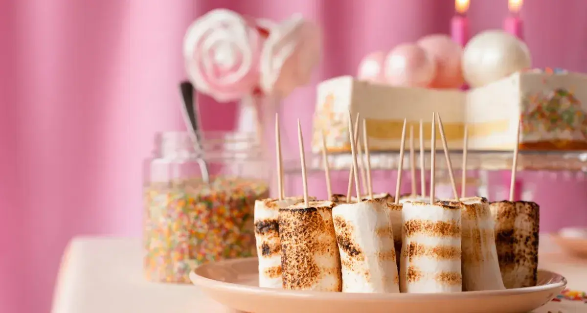 Rostade marshmallows på spett, perfekt för roliga recept att baka till kalaset. En burk strössel och en tårta i bakgrunden.