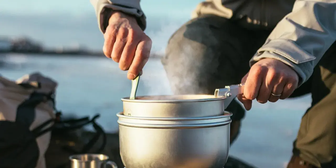 En person lagar mat utomhus, ångande kastruller och en mugg i metall antyder en lyxig matupplevelse i naturen.