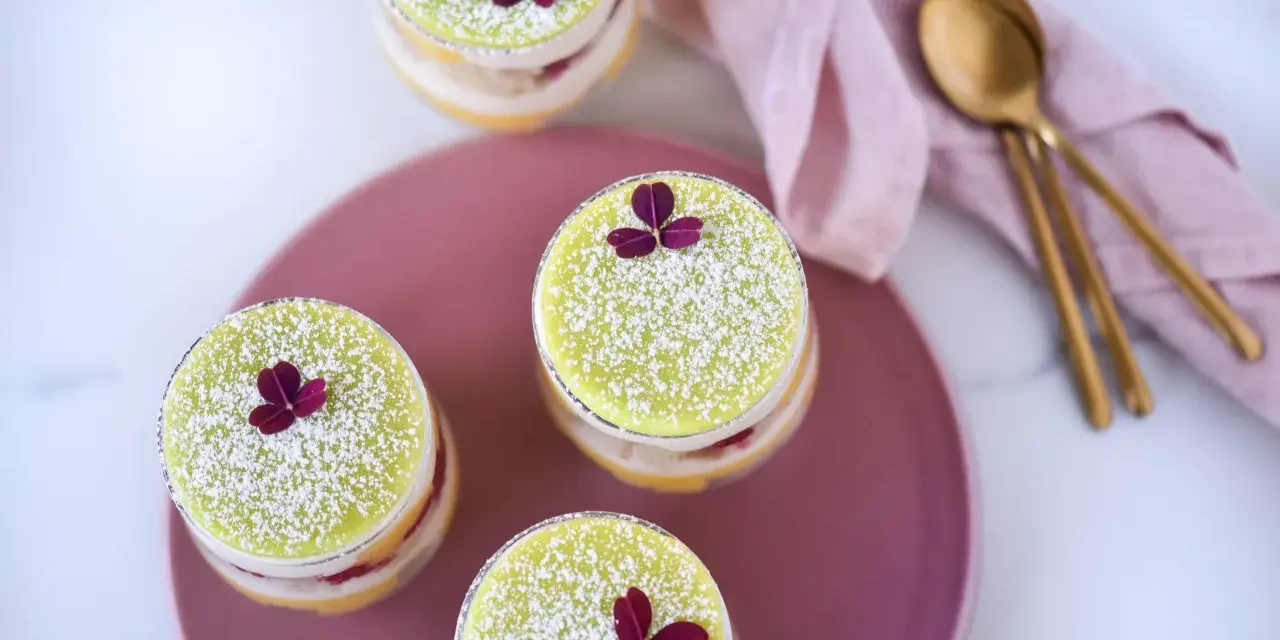 Fyra små prinsesstårtor i glas, dekorerade med florsocker och en lila blomma. En rosa duk och guldfärgade skedar ligger bredvid.