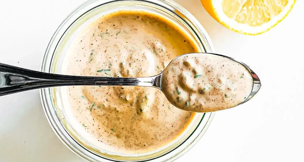 En sked krämig remouladsås med örter lyfts ur en burk. Bredvid ligger en halv citron. Perfekt för ditt remouladsås recept!