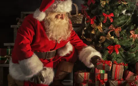 Jultomten, med sitt vita skägg och röda dräkt, sorterar julklappar vid ett dekorerat träd. Här är fakta om jultomten.