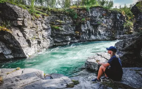 En kvinna sitter vid en forsande turkos flod omgiven av klippor. En plats för unika upplevelser i naturen.