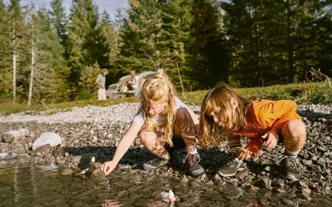Två barn leker med pappersbåtar vid en sjö, kanske en del av roliga femkampsgrenar. I bakgrunden syns ett tält och träd.