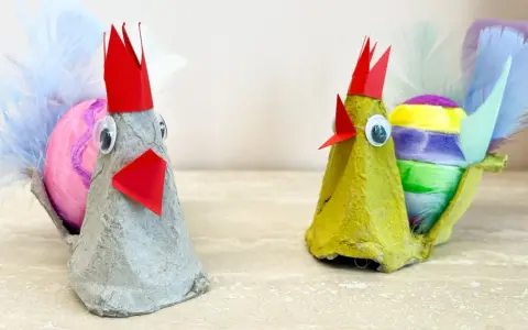Två påskkycklingar gjorda av äggkartonger, med färgglada ägg och fjädrar. Ett roligt pyssel för påsk barn.