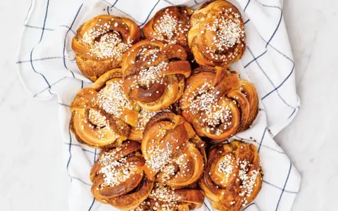En hög med gyllene kanelbullar, toppade med pärlsocker, ligger på en vit handduk med blå rutor. Camilla Hamid skulle vara stolt över dessa kanelbullar.