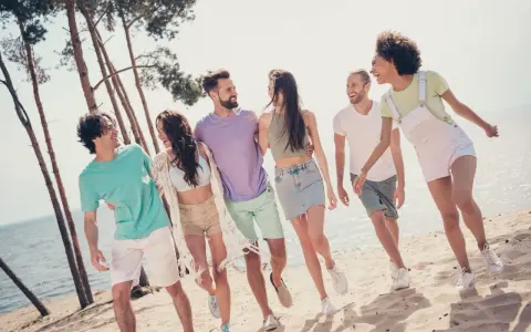 En grupp glada vänner på stranden, en riktig tipspromenad med twist. De skrattar och går längs vattnet bland tallar.