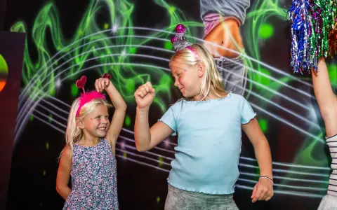 Två glada barn med diadem har roliga lekar inomhus. De dansar framför en neonfärgad bakgrund.