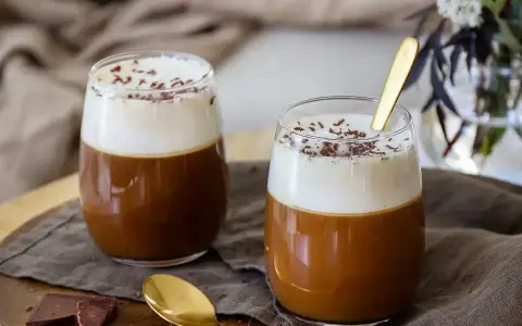 Två glas med krämig kaffe Karlsson, toppad med vispad grädde och chokladströssel. En guldig sked ligger i det ena glaset.