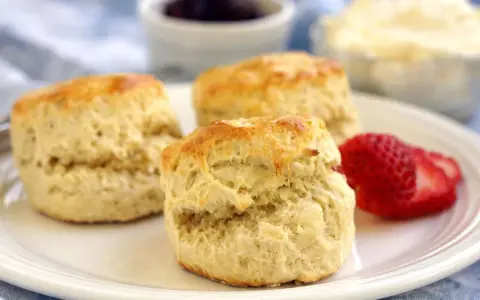 Tre gyllene engelska scones på en vit tallrik med en jordgubbe, sylt och grädde i bakgrunden.
