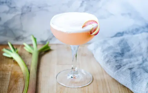 En rosa rabarber sour-cocktail med skum och en rabarberdekoration. Två rabarberstjälkar ligger bredvid.
