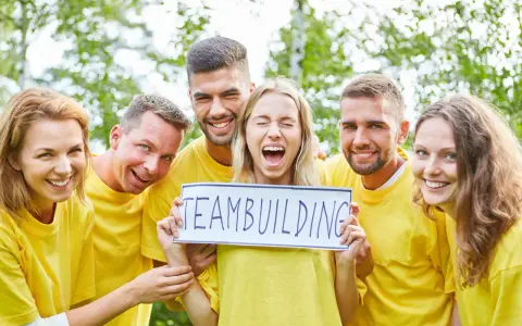 En grupp glada människor i gula tröjor håller upp en skylt med texten "TEAMBUILDING". De ser ut att ha roliga uppdrag i grupp.