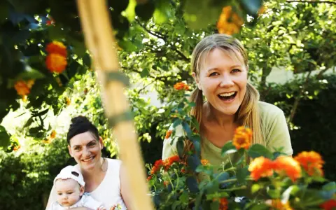 Två glada kvinnor och en bebis njuter av en solig dag utomhus, kanske under en tipspromenad med utmaningar för vuxna bland blommor och grönska.
