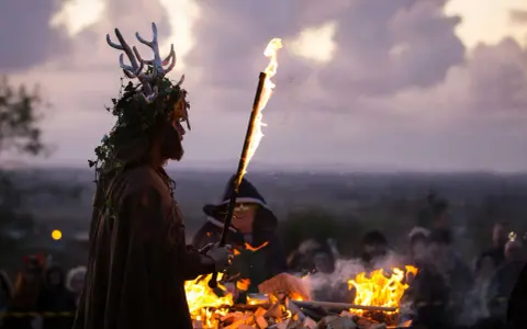 En person med hjorthorn och lövverk håller en fackla vid en eld. En mystisk stämning under halloween i Irland.