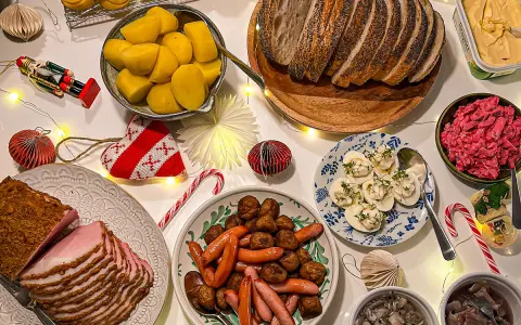Julbord med jullekar vid bordet: skinka, köttbullar, prinskorv, sill, ägg och bröd. Julpynt och ljusslingor skapar stämning.