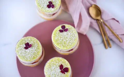 Fyra små prinsesstårtor i glas, dekorerade med florsocker och en lila blomma. En rosa duk och guldfärgade skedar ligger bredvid.