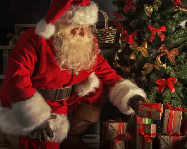 Jultomten, med sitt vita skägg och röda dräkt, sorterar julklappar vid ett dekorerat träd. Här är fakta om jultomten.