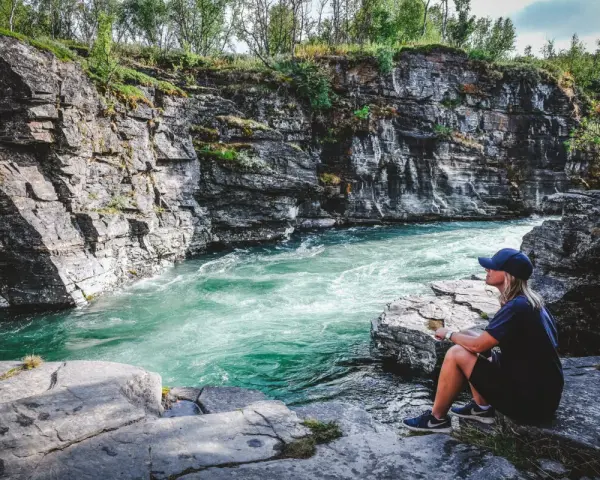 En kvinna sitter vid en forsande turkos flod omgiven av klippor. En plats för unika upplevelser i naturen.