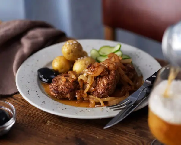En tallrik med pannbiff, stekt lök, kokt potatis, gurka och en sked med oliver. Ett glas öl hälls upp i bakgrunden. Ett klassiskt pannbiff recept.